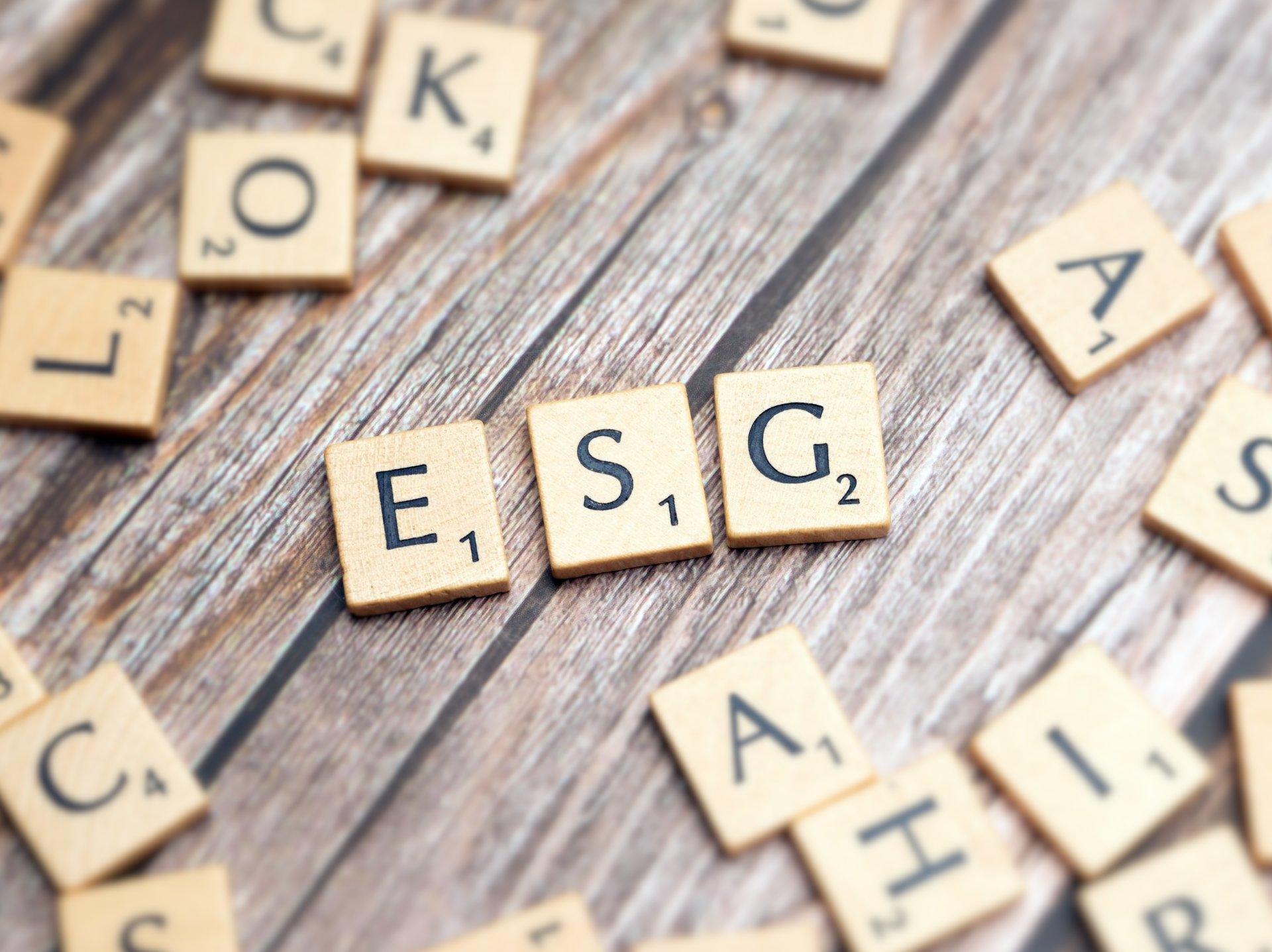 Scrabble tiles arranged on a wooden surface, with the letters 'E', 'S', and 'G' prominently displayed in the center, representing ESG (Environmental, Social, and Governance) reporting. Other scattered tiles with random letters are visible in the background.